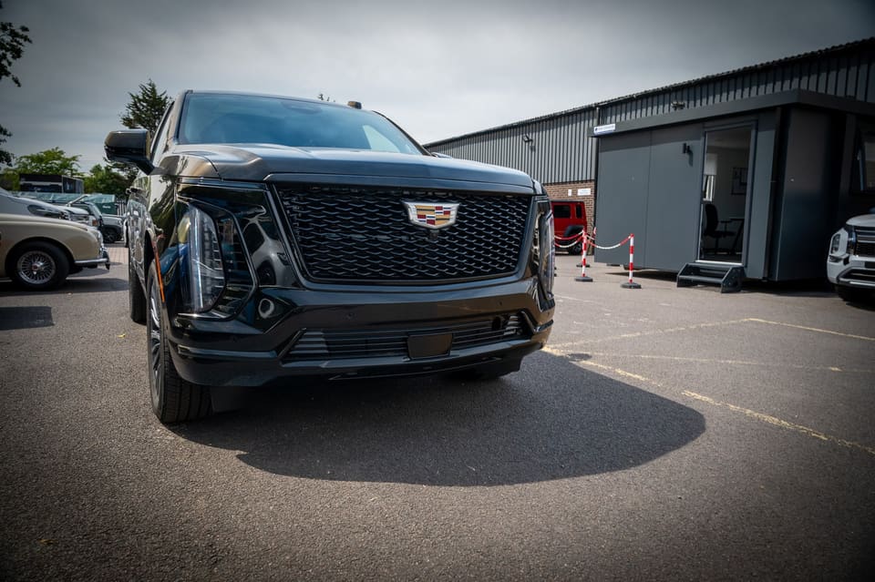 Front view of a black Cadillac Escalade parked in a yard with a warehouse building behind.