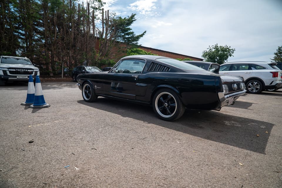 A classic Ford Mustang fastback parked in a gravel lot next to traffic cones, with other cars behind it.